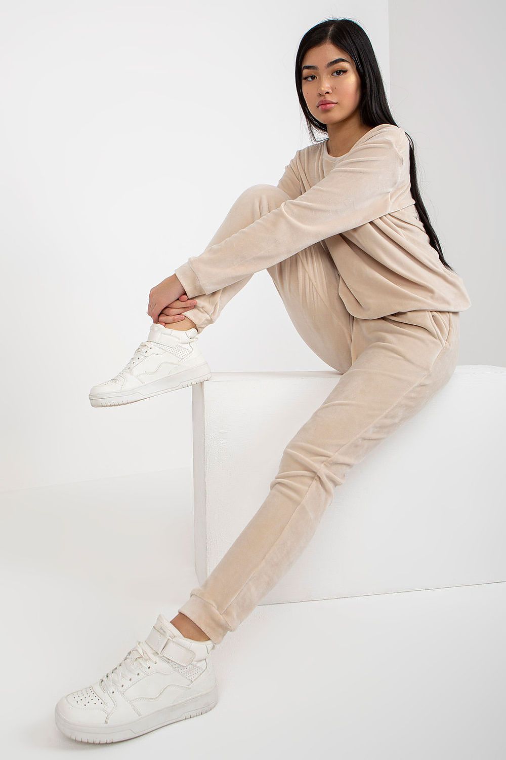 Elegant model showcasing a luxurious velour blouse and pants set by Rue Paris, styled in a soft beige tone, featuring a relaxed fit and paired with chic white sneakers against a minimalistic background.
