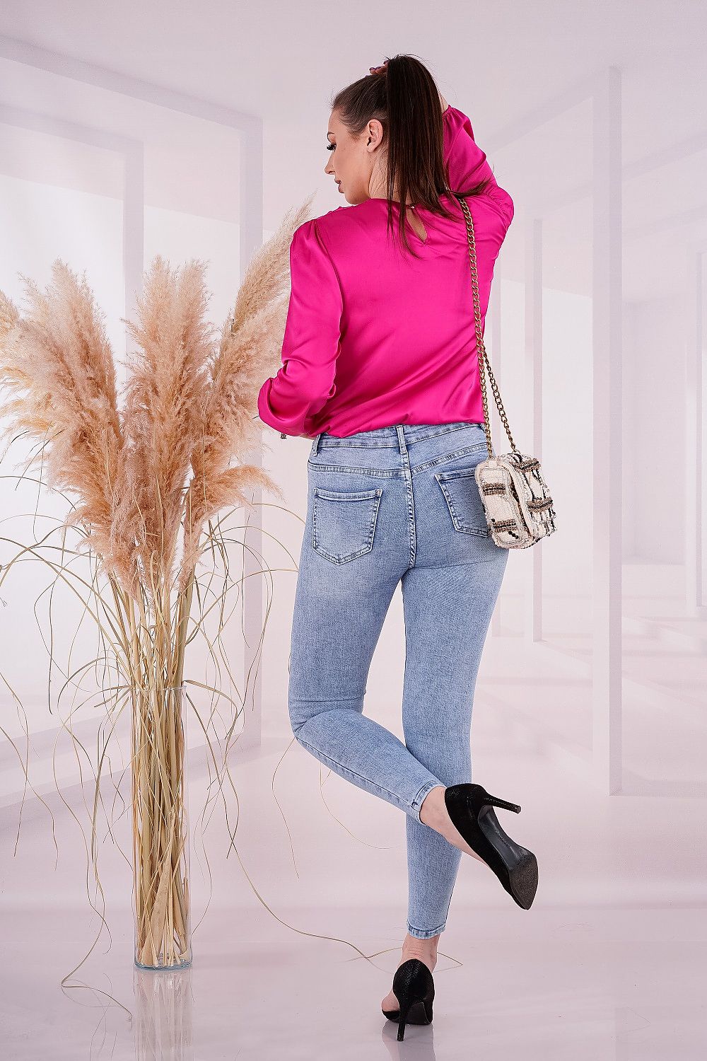 Back view of a model wearing the sophisticated Merribel satin long sleeve blouse in pink, elegantly styled with fitted denim and a stylish handbag, set against a modern, airy interior for a trendy look.
