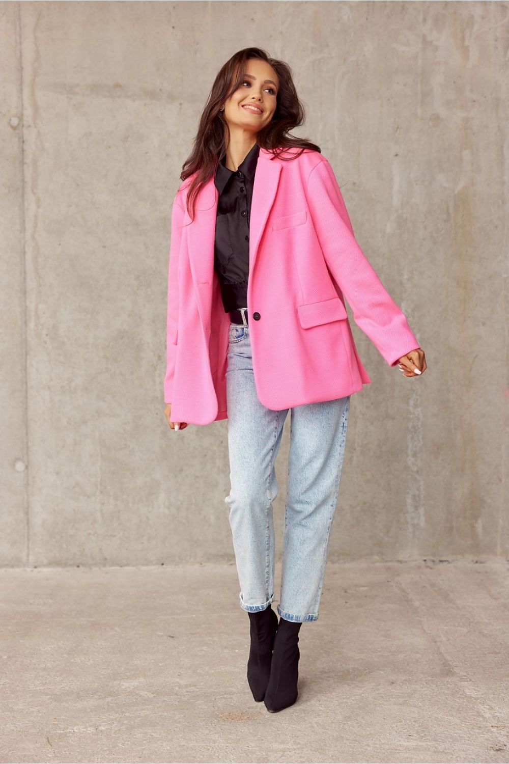 A chic model showcasing the Roco Fashion elegant voluminous pink jacket with decorative collar, styled with a black shirt and light blue jeans, exuding a sophisticated urban look against a minimalist concrete backdrop.