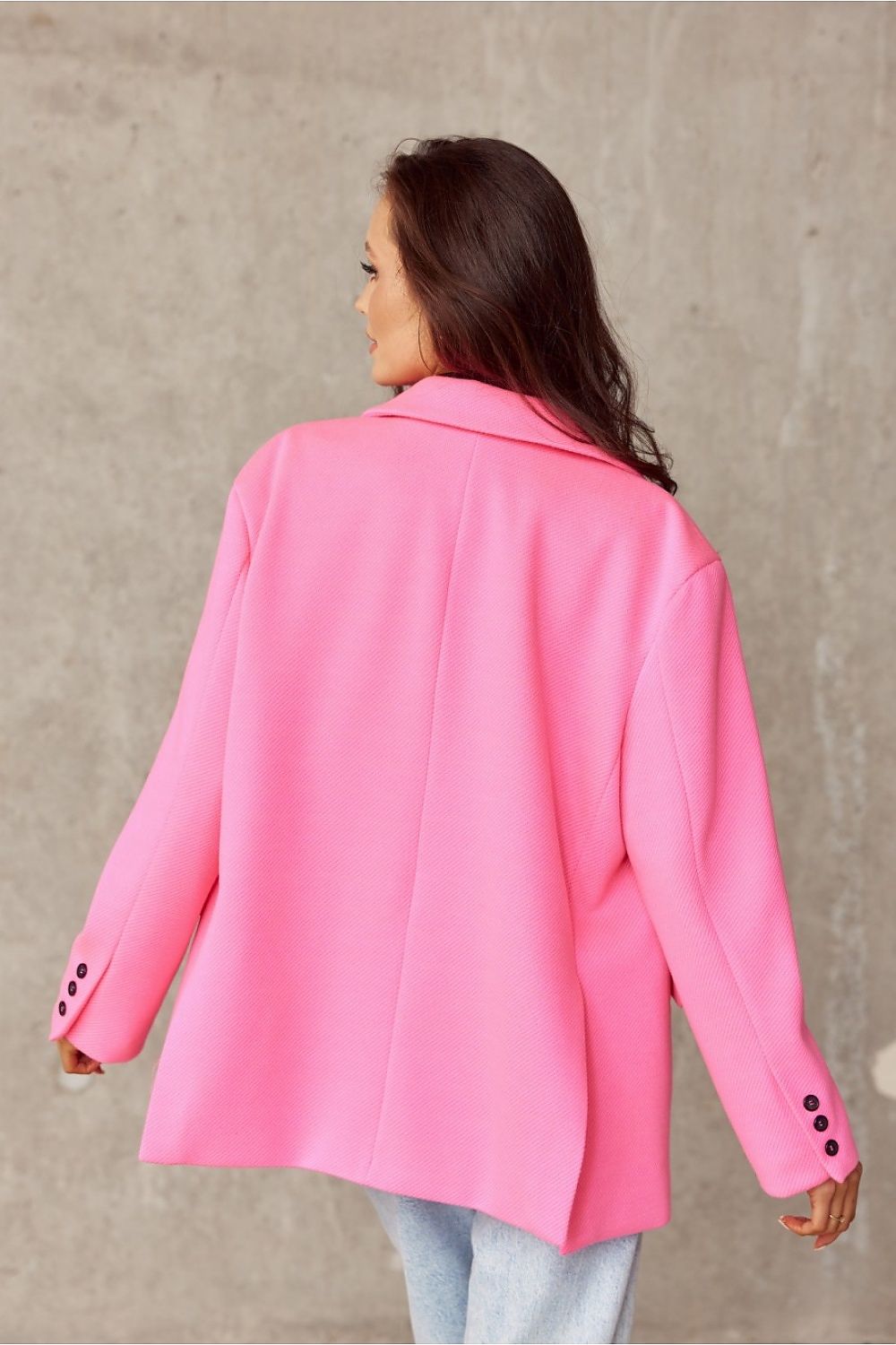 Model displaying the back view of the voluminous pink jacket by Roco Fashion, highlighting its loose fit and decorative collar, paired effortlessly with casual jeans, set against a modern concrete wall.