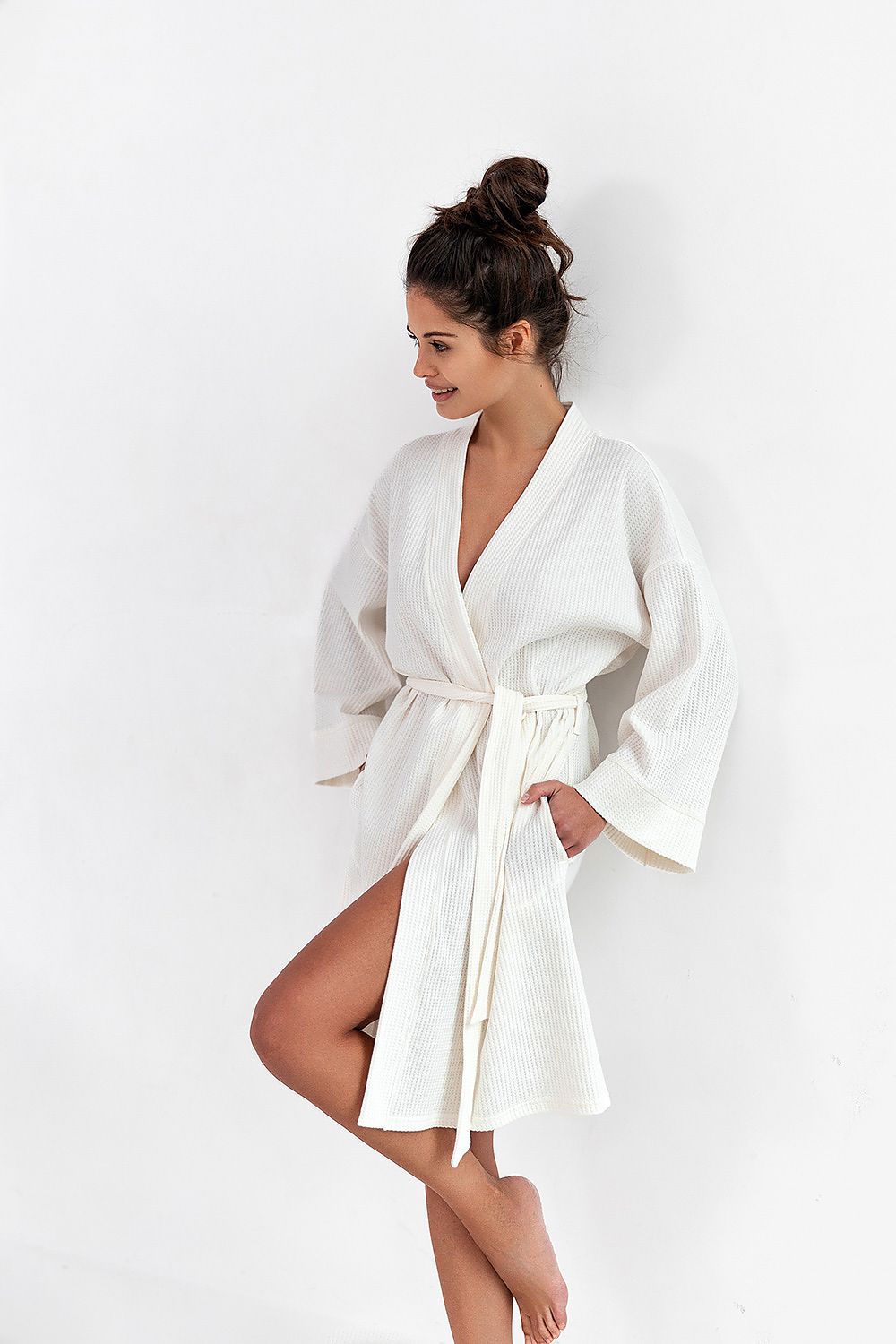Elegant woman modelling a luxury cotton bathrobe with shawl collar and functional pockets by Sensis, featuring a stylish belted waist, against a minimalistic white background.