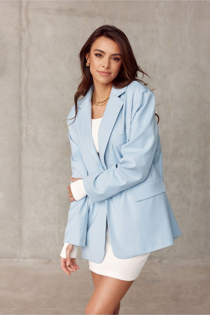 Side view of a heroine wearing a voluminous light blue jacket by Roco Fashion, featuring sophisticated detailing, perfectly complemented by a white dress.