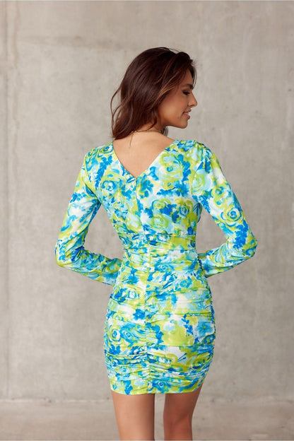 Back view of an elegant woman in a fitted mini dress by Roco Fashion with a striking floral print in blue and green. The dress boasts a V neckline and long sleeves, emphasizing a glamorous silhouette, perfect for evening occasions.