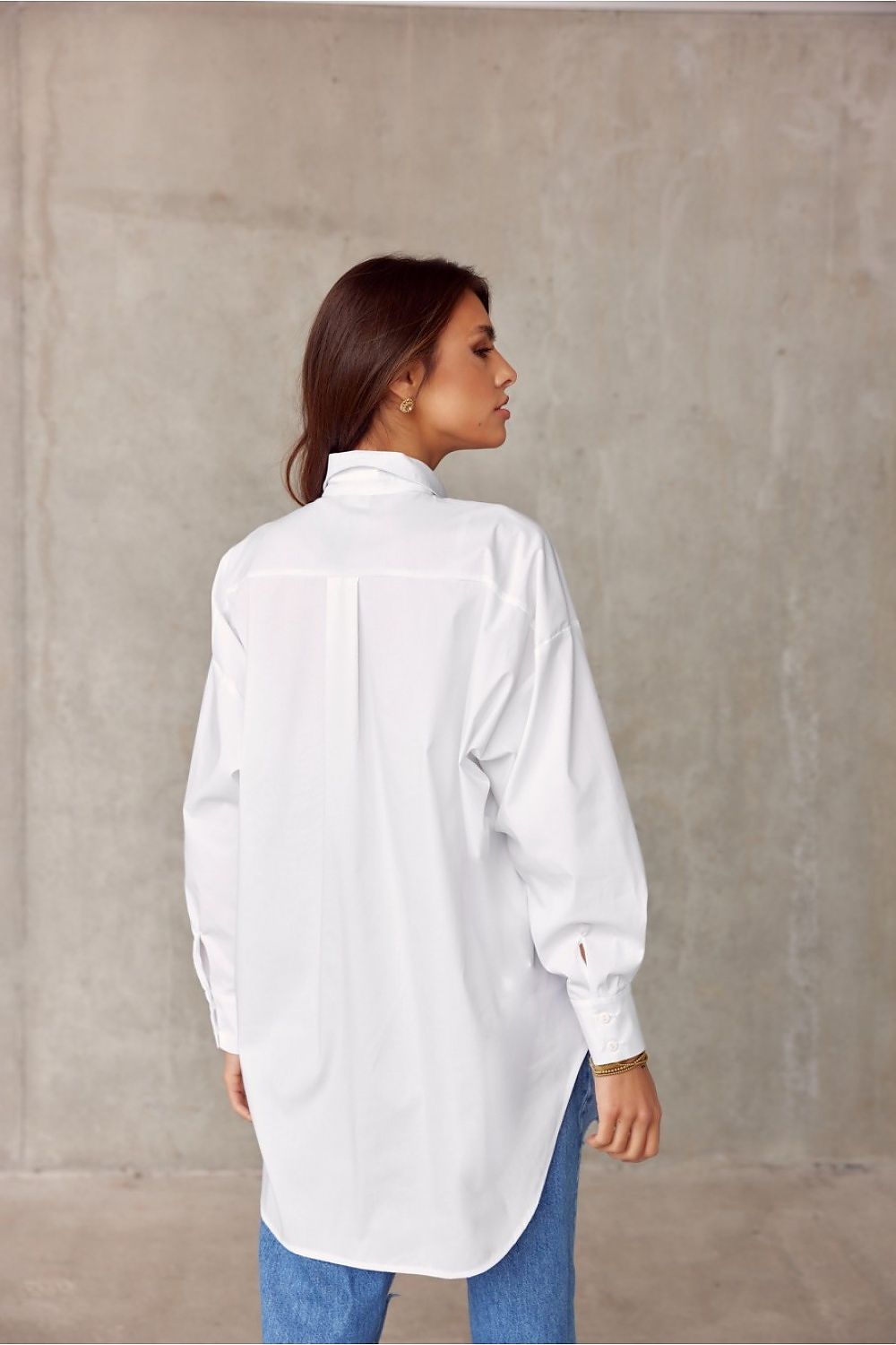 Elegant Roco Fashion women's oversized long sleeve shirt, featuring an asymmetrical hem, displayed from the back against a minimalist concrete backdrop.