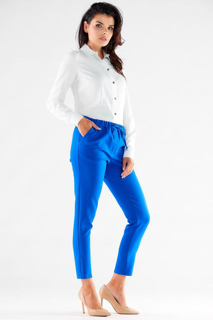 Stylish side view of a model in bright blue casual trousers by Awama, featuring a comfortable elastic waist, paired with a white buttoned shirt and pointed nude heels for a polished chic look.