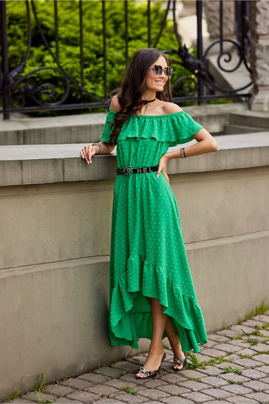 An elegant woman wearing a vibrant green asymmetrical maxi dress with ruffles and uncovered shoulders by Roco Fashion. The dress features a stylish belt and is set against a scenic background, showcasing its luxurious design and versatility for various occasions.