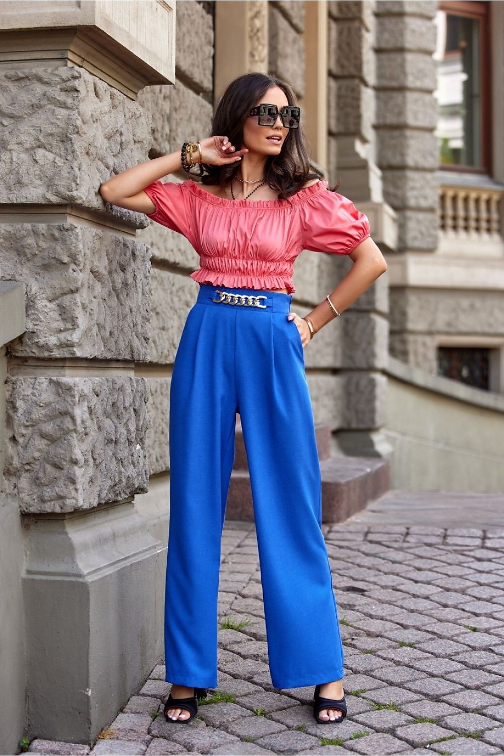 Fashionista in chic blue bell-bottom trousers by Roco Fashion, complemented by a trendy pink off-shoulder blouse and oversized sunglasses, epitomising summer elegance.