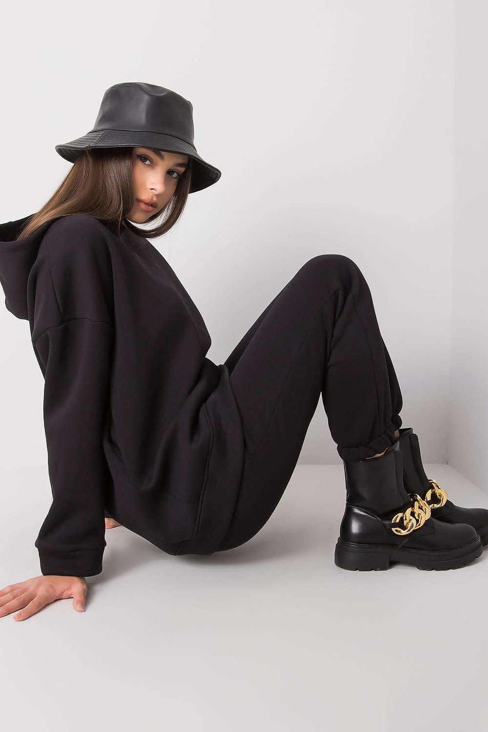 Sophisticated look of a model seated in the luxurious BFG cotton-blend sweatshirt set, featuring a hood and elastic waist pants, complemented by stylish black boots and a fashionable bucket hat.
