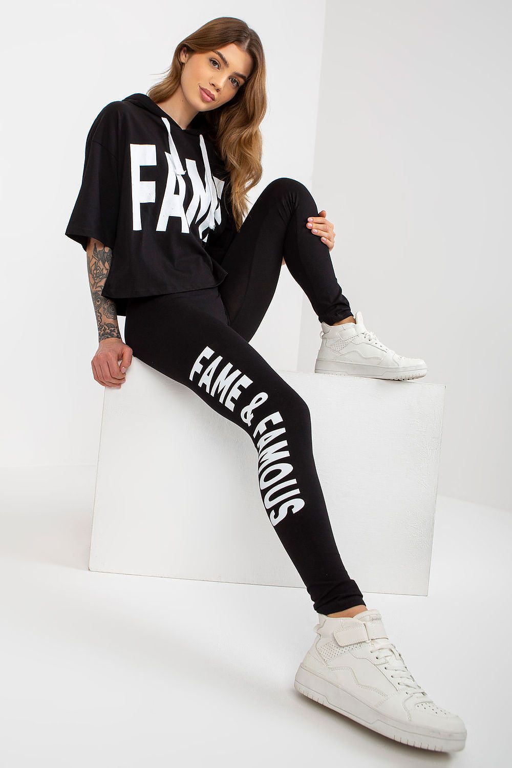 Trendy Ex Moda cotton sweatshirt and leggings set showcased by a model in black, featuring a cropped hoodie and stylish white inscriptions, complemented by fashionable high-top sneakers.