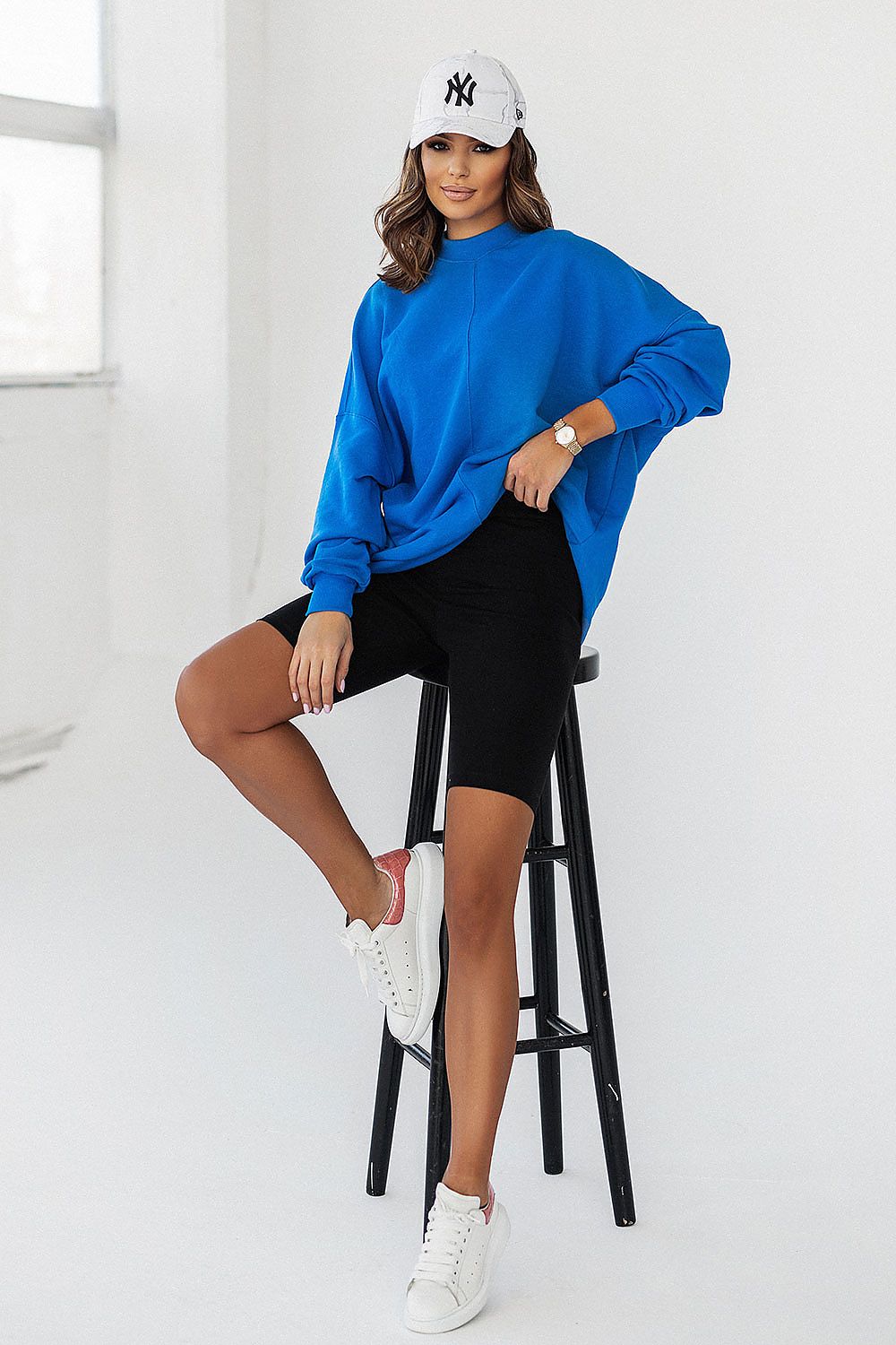 Model wearing IVON luxury blue sweatshirt, showcasing a relaxed silhouette, complemented by black bike shorts and sneakers while seated.