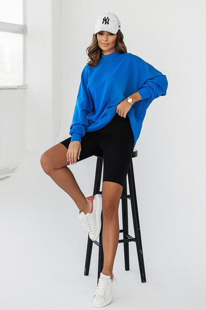 Model wearing IVON luxury blue sweatshirt, showcasing a relaxed silhouette, complemented by black bike shorts and sneakers while seated.