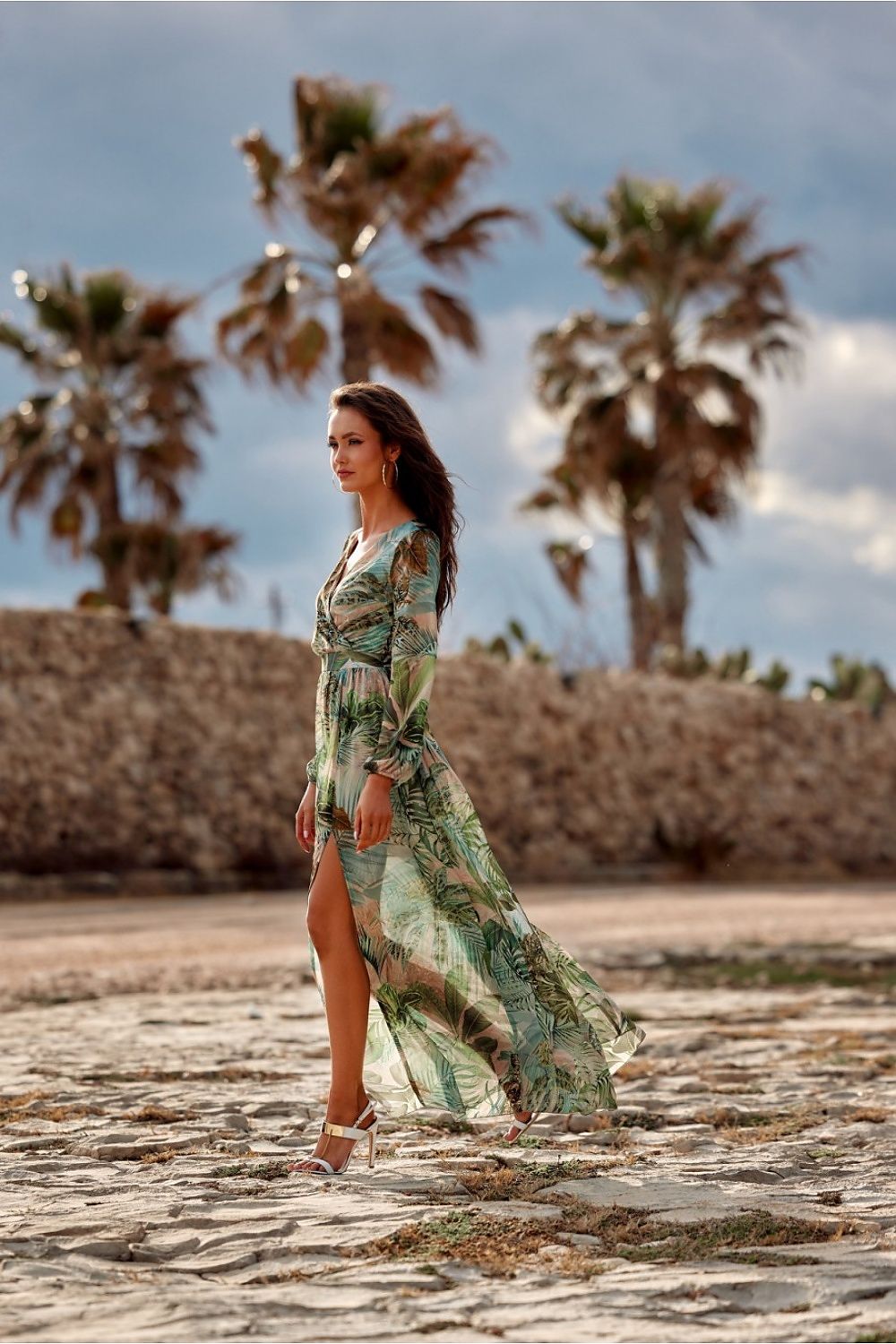 Elegant model wearing a tropical green maxi dress by Roco Fashion, featuring a thigh-high slit, amidst palm trees, showcasing a stylish summer look for weddings.