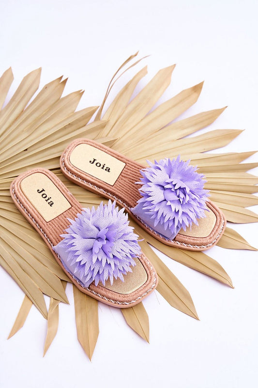A pair of luxurious women's eco-suede flip-flops by Step in Style, elegantly displayed with decorative purple flower accents against a backdrop of tropical palm leaves.