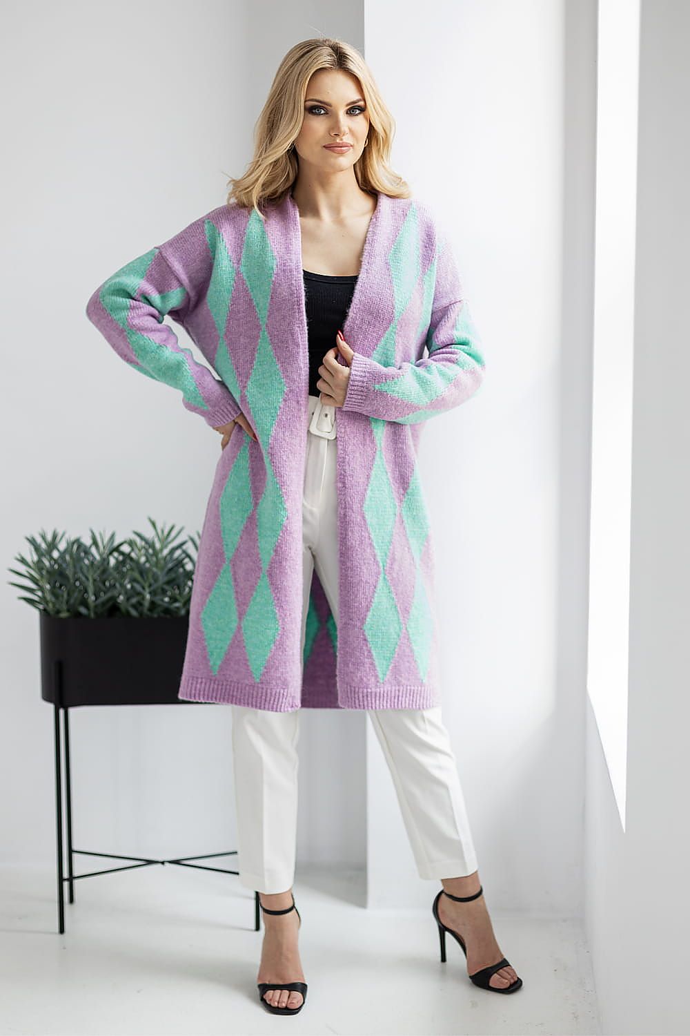 Sophisticated pose in a purple and teal geometric pattern cardigan by PeeKaBoo, paired with white trousers and strappy heels, radiating elegance and style.