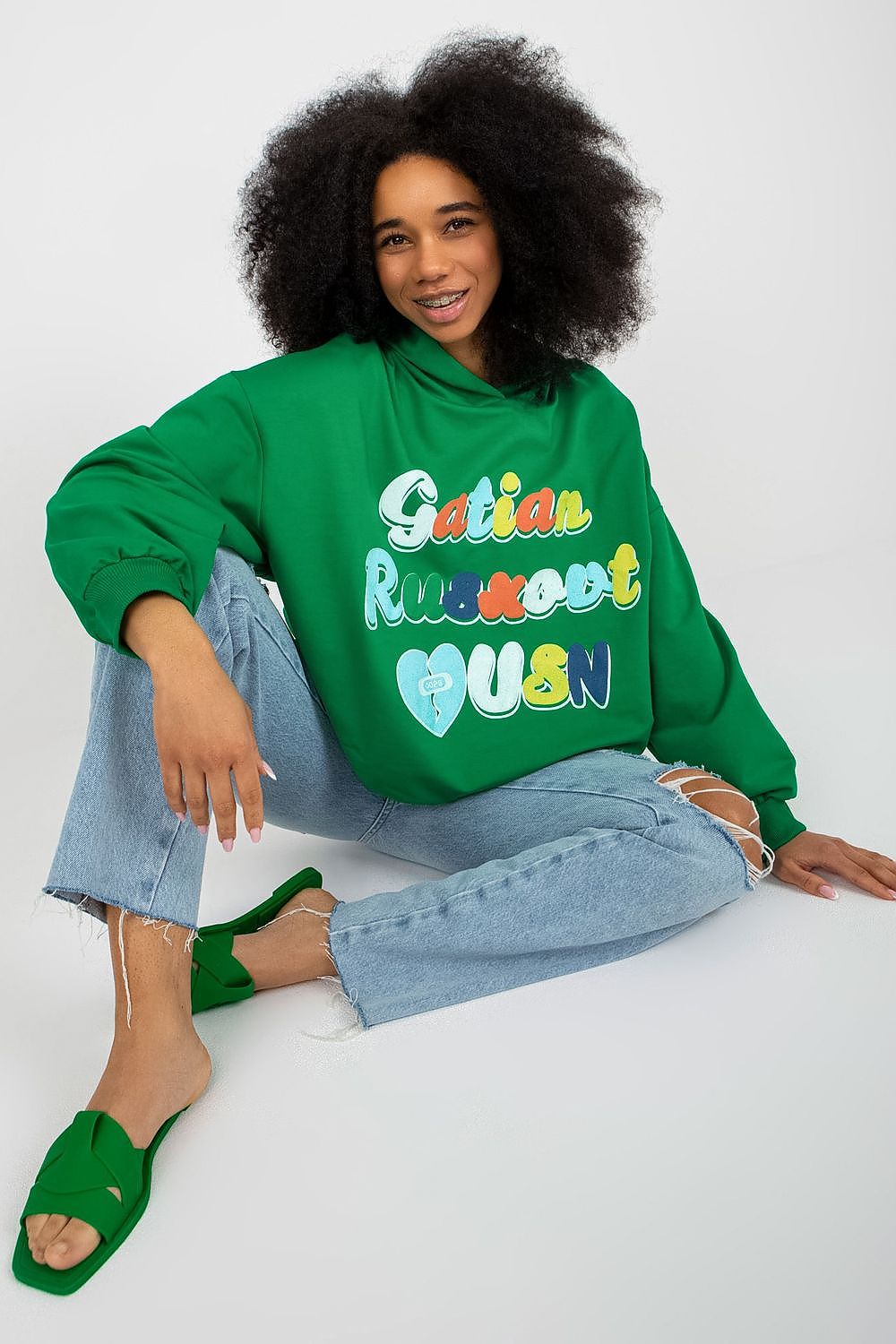 Trendy green women’s hooded sweatshirt styled casually with distressed jeans, highlighting the playful front inscription, available from Fancy.