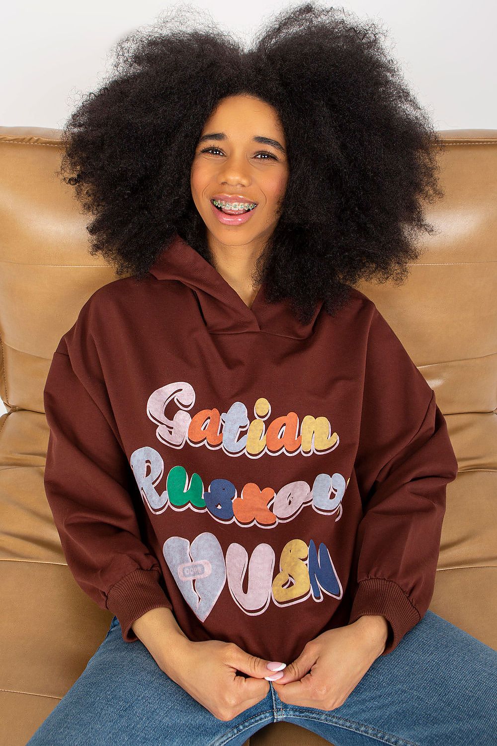 Luxurious women's hooded sweatshirt by Fancy featuring a vibrant decorative front inscription, model with curly hair sitting on a sofa.