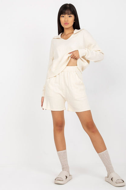 A stylish model wearing a luxurious cream 2-piece set by EM, featuring a loose sweater and shorts combination, paired with contemporary accessories for a chic look.