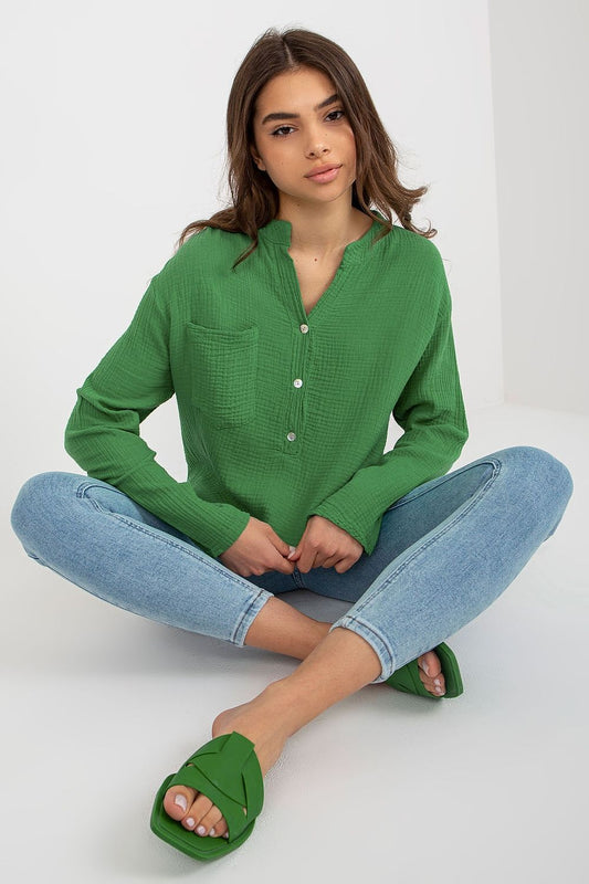 Elegant model seated wearing an Och Bella loose fit muslin blouse in green, showcasing a v-neck design and front pocket, paired with denim, exuding luxury fashion.