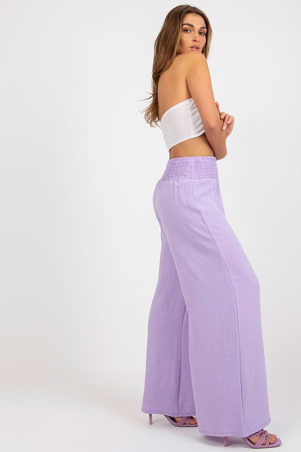 Luxury high-waisted women's trousers by Italy Moda. Features long seamed design with decorative creases, showcased on a model wearing a stylish white crop top and vibrant lilac trousers, perfect for elegant occasions.