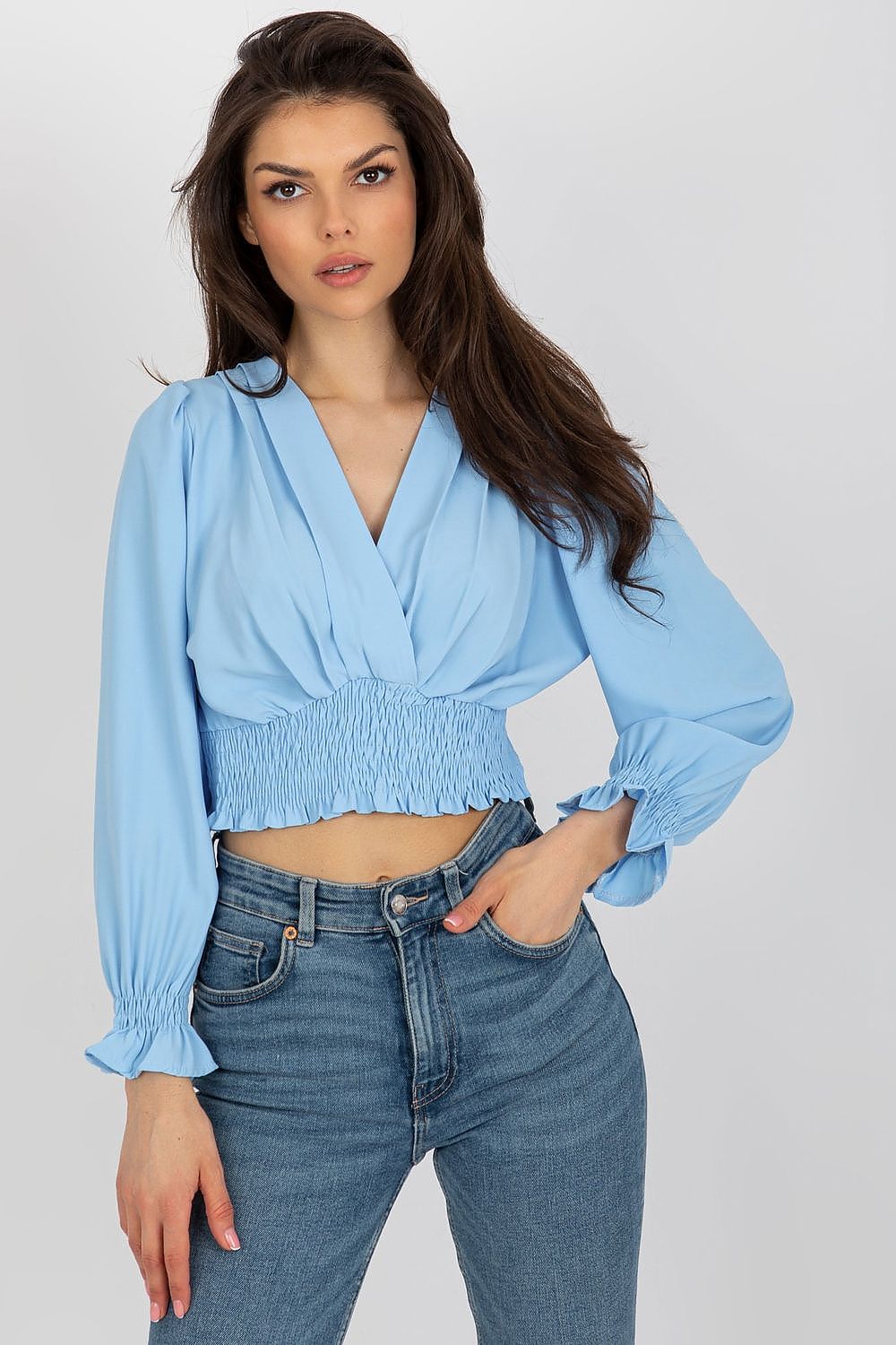 Elegant model posing in a light blue long sleeve blouse with a trendy envelope neckline and smocked waist from Italy Moda, complemented by high-waisted jeans.