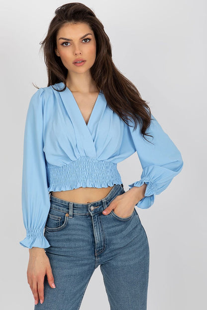 Elegant model posing in a light blue long sleeve blouse with a trendy envelope neckline and smocked waist from Italy Moda, complemented by high-waisted jeans.