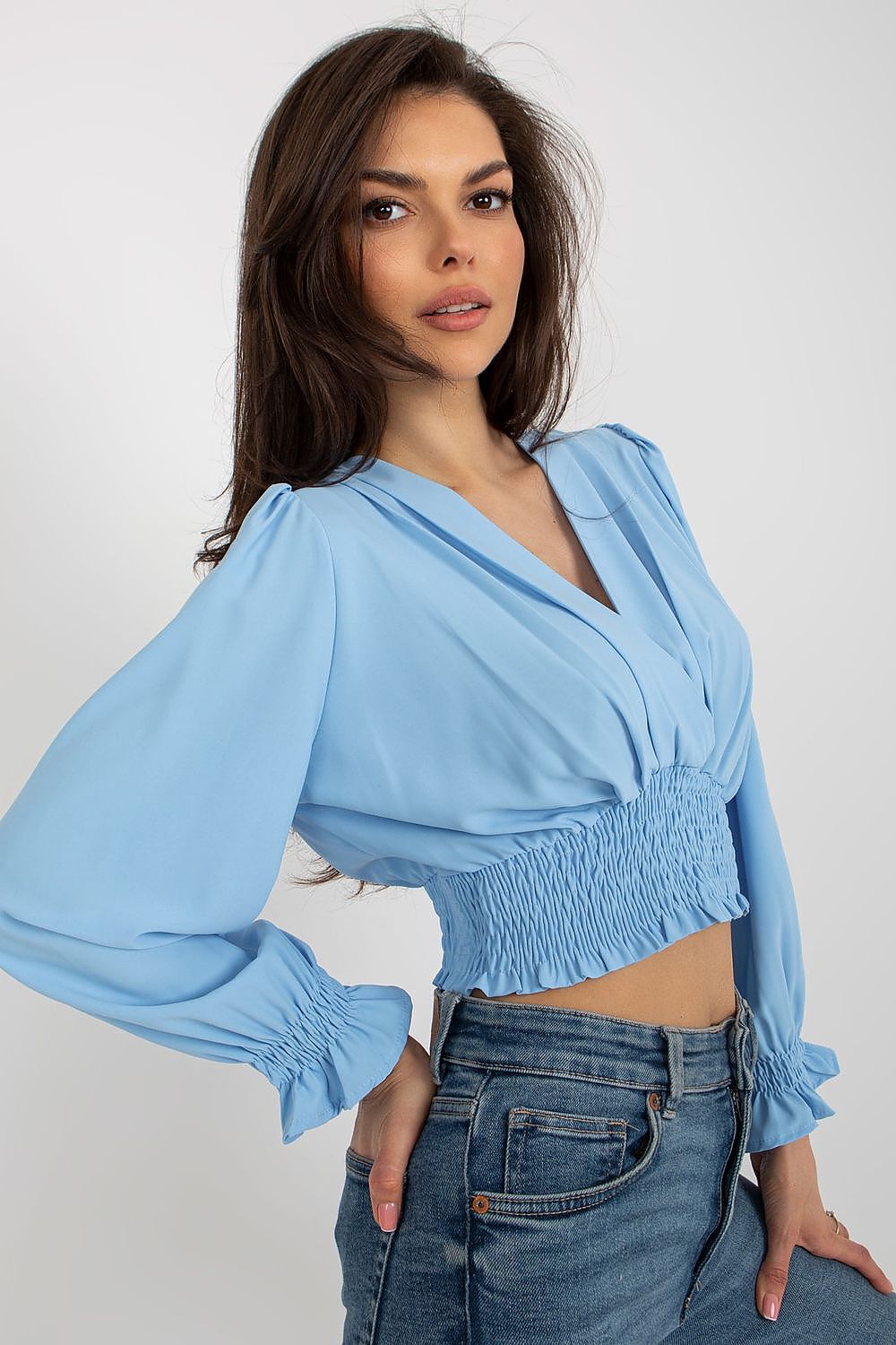 Stylish close-up of a model in a light blue blouse featuring an envelope neckline and smocked design at the waist from Italy Moda, perfectly matched with denim jeans.